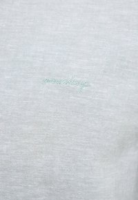 Jasnoszara koszula z tkaniny z delikatną teksturą, ozdobiona haftowanym logo "mustang" w miękkim zielonym kolorze z przodu.