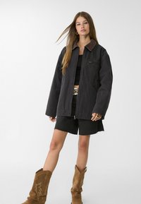 Zwarte oversized jas met een bruine corduroy kraag, gecombineerd met een zwarte tanktop en denim shorts. Model draagt bruine cowboylaarzen.
