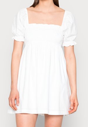 Frau trägt ein kurzes weißes Kleid mit Puffärmeln und quadratischem Ausschnitt, steht vor einem schlichten hellen Hintergrund.