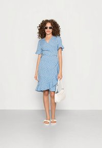 Žena s kovrčavom kosom, u sunčanim naočalama, plavoj cvjetnoj haljini na preklop, bijelim sandalama i bijelom torbicom, stoji protiv jednostavne pozadine.