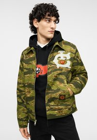 Πράσινο μπουφάν με πατρόν καμουφλάζ και φερμουάρ, με λευκές και κόκκινες γραφικές λεπτομέρειες, συνοδευόμενο από μαύρη κουκούλα.