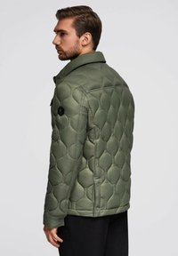 Uomo con capelli corti castani e barba, che indossa una giacca verde oliva trapuntata con polsini con bottoni, in piedi di lato su uno sfondo semplice.