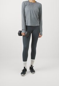 Harmaa pitkähiha-paita, kuvioidut tummanharmaat legginsit, mustat urheilukengät valkoisilla pohjilla ja musta käsipaino kädessä.