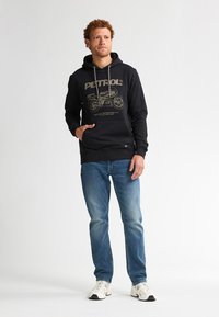 Felpa nera con grafica "PETROL" in color sabbia, tasca a marsupio frontale, abbinata a jeans denim blu e sneakers bianche. Sfondo neutro.