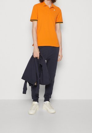 Personne portant un polo orange à manches courtes, un pantalon de jogging bleu marine, des baskets blanches, tenant un sweat à capuche bleu marine plié, sur un fond clair uni.