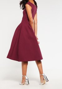 Abito borgogna off-the-shoulder con busto aderente, gonna plissettata e tessuto testurizzato. Sandali con tacco alto argentati completano il look.