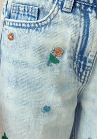 Lyseblåt denimstof med små broderede orange, blå og ferskenfarvede blomster nær en forlomme og bæltestrop.