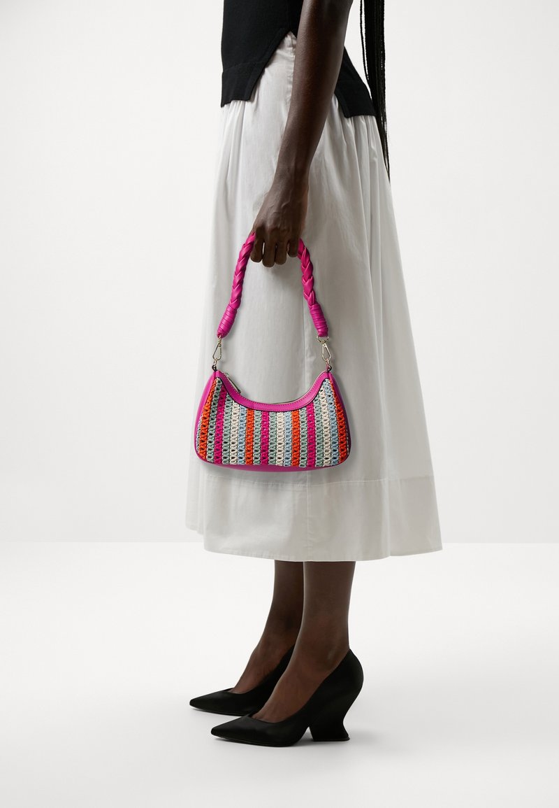 Femme portant une jupe blanche et un haut noir, tenant un petit sac à main tissé rayé avec une poignée tressée rose, portant des talons pointus noirs.