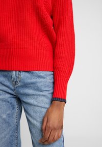 Rød ribstrikket sweater med blokmønster og mørkeblå detaljer på ærmekanterne, kombineret med lyseblå denimjeans.