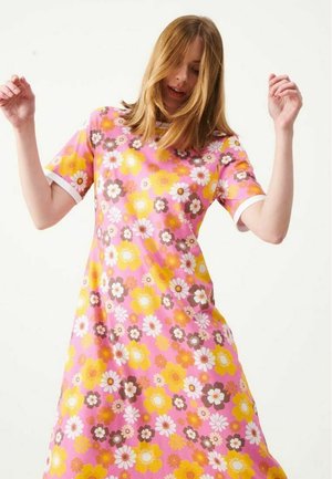 Kvinde med skulderlangt hår iført en lyserød kjole dækket af gule, hvide og brune blomsterprint, der løfter begge hænder let.