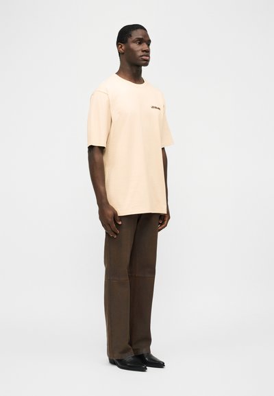 Άνδρας στέκεται φορώντας μια μπεζ φαρδιά μπλούζα, καφέ σκούρο ευθείες παντελόνια και μαύρα παπούτσια με μυτερά δάχτυλα, σε απλό φωτεινό φόντο.