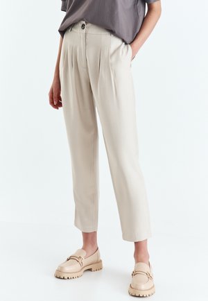Beżowe plisowane spodnie z wysokim stanem noszone z taupe topem i masywnymi beżowymi mokasynami, modelka stoi z jedną ręką w kieszeni na białym tle.