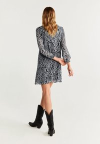 Vestido negro y azul marino con estampado de cachemira, mangas largas transparentes, puños acampanados y un corte suelto. Llevado con botas de cowboy negras.