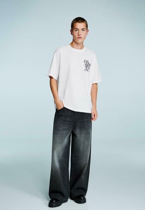 Hvid bomulds t-shirt med et lilla grafisk logo på venstre bryst, kombineret med brede, blege sorte denim jeans og sorte klodsede sko.