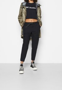 Kvinna i svart Columbia crop top, olivgr ön regnjacka, svarta cargobyxor med bälte och svart-vita högskor.