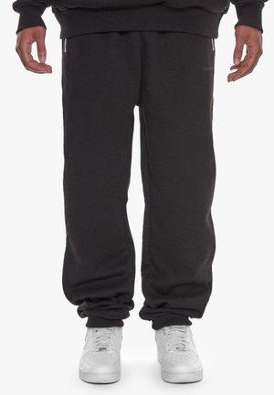 Persoon die zwarte joggingbroek en witte sneakers draagt, staat met ontspannen armen langs het lichaam op een witte achtergrond.