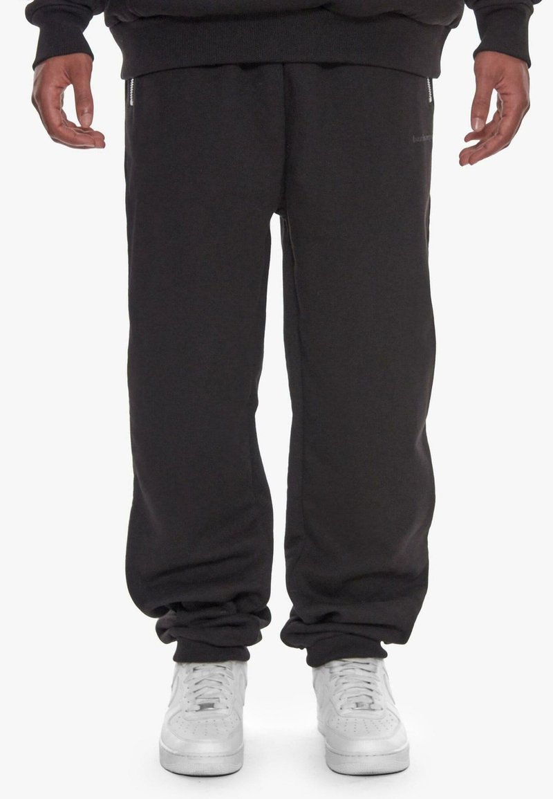 Personne portant un pantalon de survêtement noir et des baskets blanches, debout, les bras détendus le long du corps sur un fond blanc.