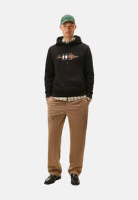 Sudadera negra con logo, camisa de cuadros debajo, pantalones marrones a rayas y zapatos negros. Modelo con una gorra verde, manos en los bolsillos.