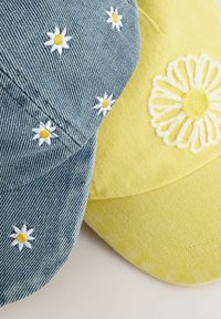 Denim sapka fehér és sárga hímzett százszorszépekkel, mellette egy sárga sapka, amelyen egy nagy fehér százszorszép minta található. Puha textúrák, laza stílus.