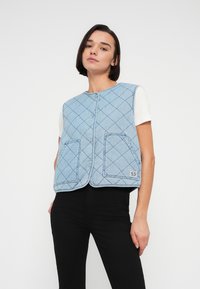 Αμάνικο τζιν μπουφάν σε ανοιχτό μπλε χρώμα με ρομβοειδές σχέδιο, κουμπιά με τυφλά κουμπώματα και δύο μπροστινές τσέπες. Φορεμένο πάνω από λευκό κοντομάνικο πουκάμισο.