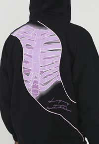 Black hoodie featuring a large, stylized purple ribcage graphic on the back, accented with a contrasting white outline and text.