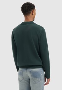 Donkergroene trui met lange mouwen, een polokraag en geribbelde zoom, gecombineerd met lichtblauwe jeans met achterzakken en een vervaagde textuur.