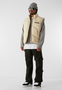 Chaleco acolchado beige con diseño de acolchado, suéter gris, camiseta blanca debajo, pantalones cargo negros y zapatillas deportivas con patrón.