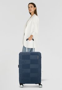 Valigia in policarbonato testurizzato blu navy con un motivo geometrico, dotata di due ruote e manico retrattile. Si trova in posizione eretta accanto a una persona in giacca.