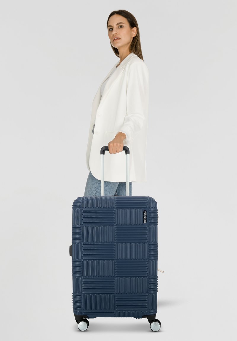 Valigia in policarbonato testurizzato blu navy con un motivo geometrico, dotata di due ruote e manico retrattile. Si trova in posizione eretta accanto a una persona in giacca.