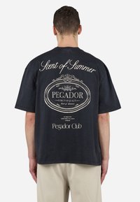 Tricou oversized negru cu text și grafică imprimate în alb pe spate, inclusiv „Aromă de Vară” și „Clubul Pegador.”