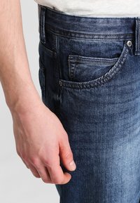 Mörkblå denimjeans har sydda fickor och en slät yta, med en hand som vilar på midjebandet för skala och sammanhang.