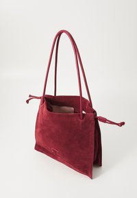 Sac à main en daim rouge avec une forme structuré, deux poignées et une fermeture à cordon. Il présente une doublure intérieure lisse et un détail de logo embossé.