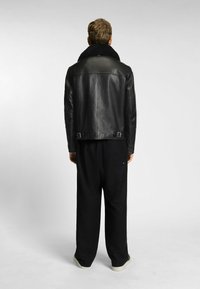 Schwarze Lederjacke mit hohem Kragen und verstellbaren Schnallen an der Taille, kombiniert mit locker sitzenden schwarzen Hosen und hellen Schuhen.