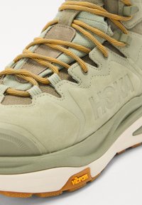 Pantof de drumeție din suede verde deschis, cu șireturi portocalii și o talpă căptușită. Prezentă un material texturat și un logo Vibram proeminent pe talpă.