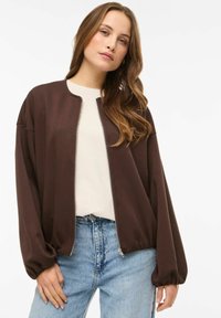 Pruun pommitajaja tükk ümara kraega, esikäepidemega ja pallivarrukatega, kantud kreemja topi kohal, ühendatud helesiniste teksadega.