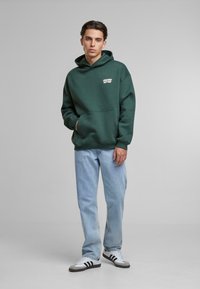 Hanorac verde cu buzunar frontal și logo alb, asortat cu blugi ușor albaștri și adidași gri cu accente negre. Design simplu și casual.
