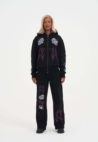 Zwarte hoodie met rits, versierd met paarse vlamgraphics en doodskopontwerpen, gecombineerd met zwarte sweatpants met vergelijkbare motieven en een relaxte pasvorm.
