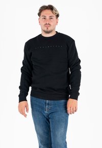 Uomo con capelli corti che indossa una felpa nera a maniche lunghe con scritte leggere e jeans blu, in piedi davanti a uno sfondo bianco.