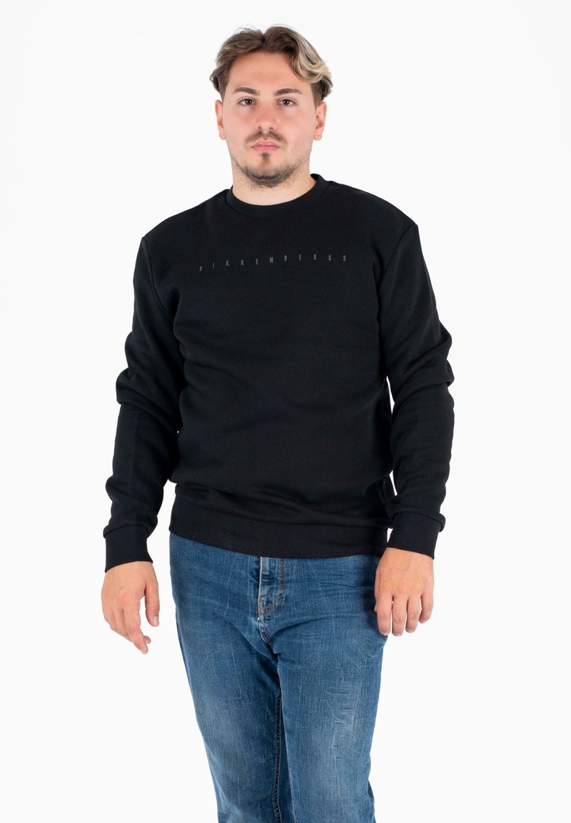 Uomo con capelli corti che indossa una felpa nera a maniche lunghe con scritte leggere e jeans blu, in piedi davanti a uno sfondo bianco.