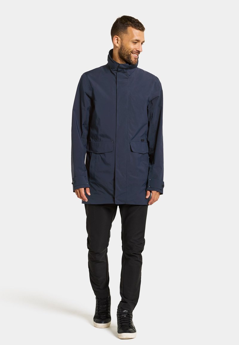 Marineblaue wasserdichte Jacke mit hohem Kragen, zwei vorderen Taschen und verstellbaren Bündchen. Getragen über schwarzen Slim-Fit-Hosen und high-top Sneakers.