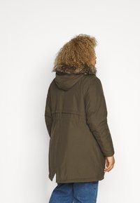 Parka de color verde oliva con capucha forrada de piel sintética, mangas largas y cintura ajustada. Presenta una abertura en la espalda para facilitar el movimiento. Textura suave.