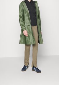 Zielony wodoodporny płaszcz przeciwdeszczowy z kapturem i zapięciem na napy z przodu, w zestawieniu z czarną koszulą zapinaną na guziki i jasnobrązowymi, dopasowanymi spodniami.