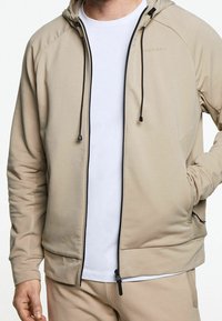 Chaqueta beige con cremallera y capucha, con herrajes y acentos negros, fabricada en un tejido suave, con mangas raglán y bolsillos frontales.