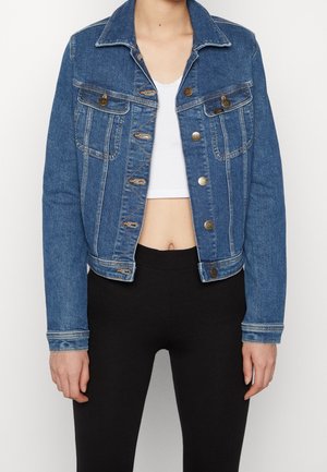 Vrouw draagt een blauwe spijkerjas over een wit cropped topje en zwarte legging, staand tegen een effen witte achtergrond.