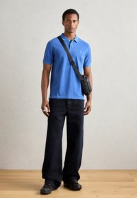 Blue polo shirt, cotton fabric, short sleeves, buttoned collar; paired with loose black jeans and black crossbody bag. Dark footwear and rings visible.