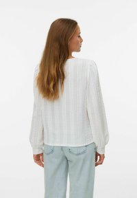 Femme aux longs cheveux bruns portant un chemisier blanc texturé à manches longues et un jean bleu clair, de dos contre un fond uni.