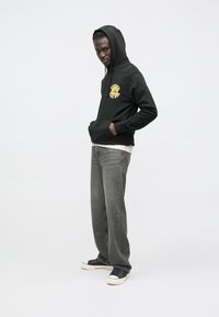 Felpa nera con cappuccio e stampa grafica raffigurante un leone giallo, abbinata a jeans grigi e scarpe da ginnastica nere. Dotata di tasca frontale e cappuccio.