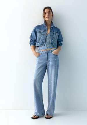 Femeie stând lângă un perete alb, purtând o jachetă scurtă din denim, top din denim, blugi evazați și sandale negre, cu mâinile în buzunare.