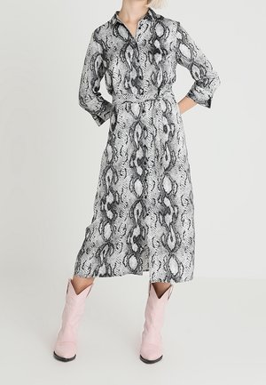 Kvinde iført en langærmet, knap-op midi-kjole med slangeprint, kombineret med lyserøde cowboystøvler, stående med hænderne på hoften.