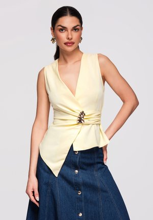 Kobieta ubrana w bladoróżowy bezrękawnik z dekoracyjną zapinką i ciemnoniebieską jeansową spódnicę na guziki, pozująca z jedną ręką na biodrze.